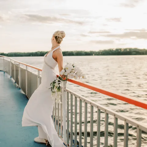 Bride in white dress holding flowers on a boat, with a scenic water view.