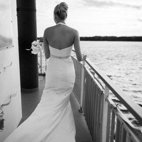 Bride in gown stands on a boat, overlooking a lake.