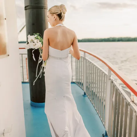Bride in white gown holding flowers on a boat deck overlooking water.