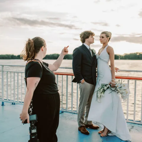 Photographer directs couple posing on a boat, bride in white gown, groom in suit, holding a bouquet.