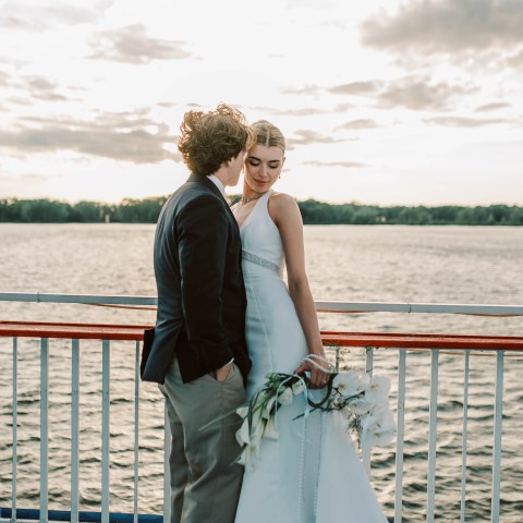 Couple on boat deck, man in suit kisses woman in white dress, holding bouquet, water and sky in background.