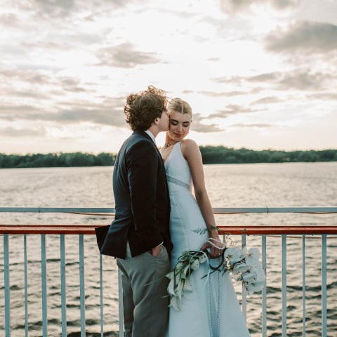 Couple in wedding attire on a boat deck by a lake under a cloudy sky.