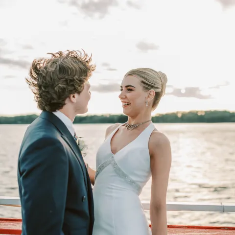 Couple in formal attire smiling by a lakeside railing at sunset.