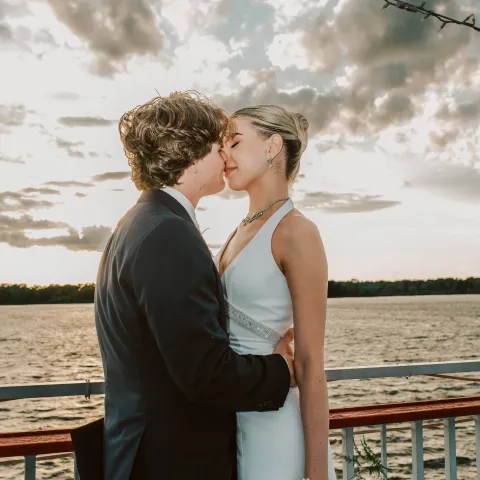 Couple in formal attire embracing on a boat at sunset.