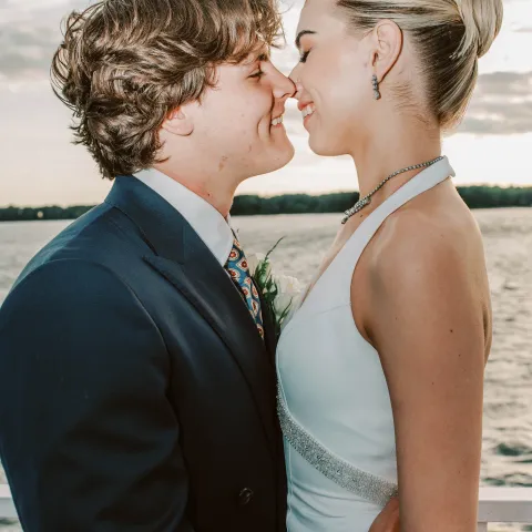 Couple in formal attire embracing by a waterfront at sunset.