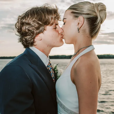A couple in formal attire kissing by a lake at sunset.