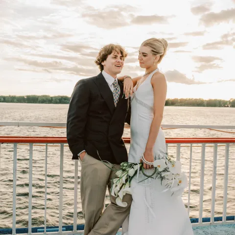 Couple in formal attire on a boat deck at sunset.