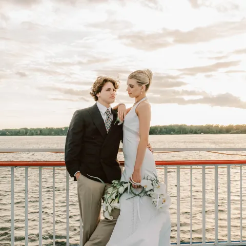 Couple posing on a boat deck, woman in a white dress holding flowers.