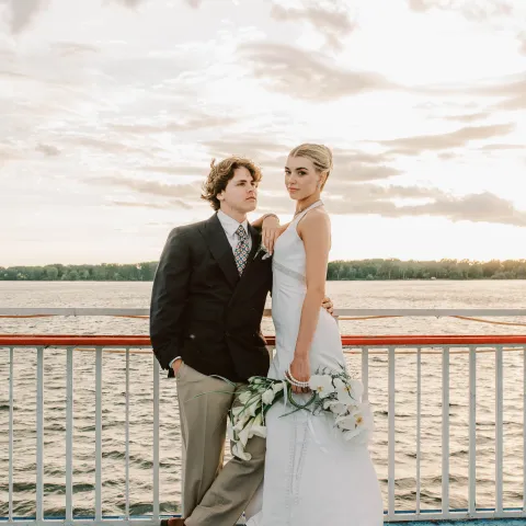 Couple in formal attire stands on a boat deck with water and cloudy sky backdrop.