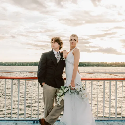 Couple on a boat deck; man in suit, woman in white dress with bouquet, water and sky in background.