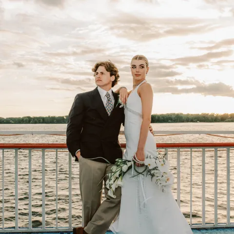 Couple in formal attire on boat deck with sunset over water.