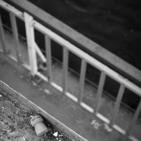 Black and white photo of a metal railing with a champagne cork on a ledge above water.