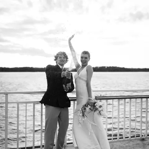 Couple celebrating on a boat with champagne, bride holds bouquet.