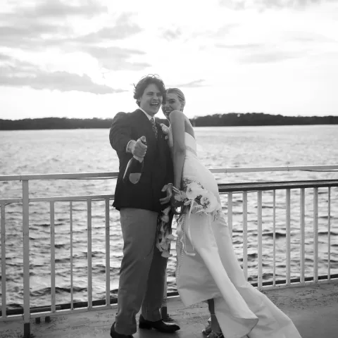 Couple in wedding attire smiling by a lake, overcast sky.
