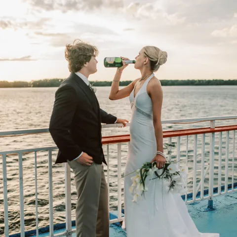 Couple on boat, woman in wedding dress drinks from bottle beside man in suit.