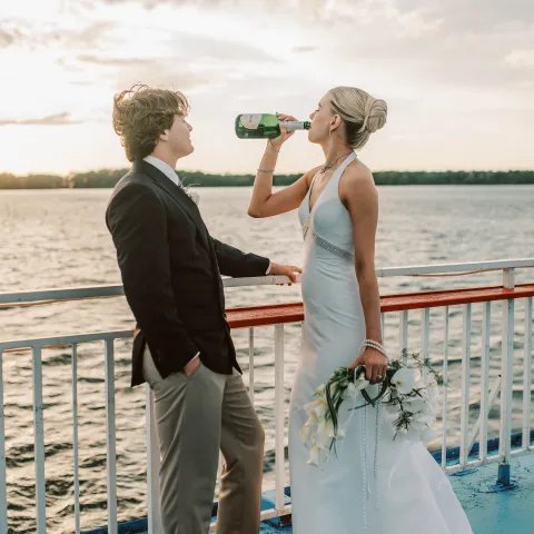 Bride drinks from bottle, groom watches on boat deck at sunset.