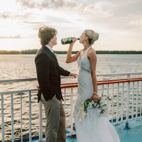 Bride drinks from bottle, standing with groom on boat deck at sunset.