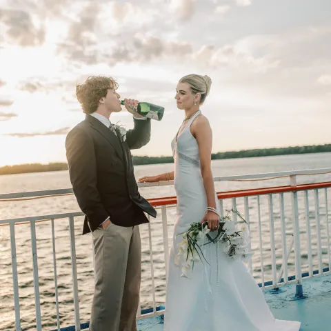 Bride and groom on a boat, groom drinking from a bottle, sunset in the background.