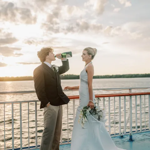 Bride holding flowers, groom drinking bottle on a boat deck at sunset.