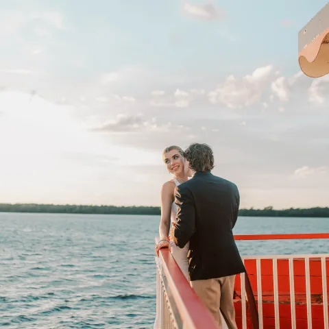 Couple on a boat, leaning on the railing, with a scenic water view.