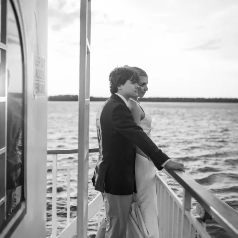 A couple in formal attire stand on a boat deck, gazing at the water.