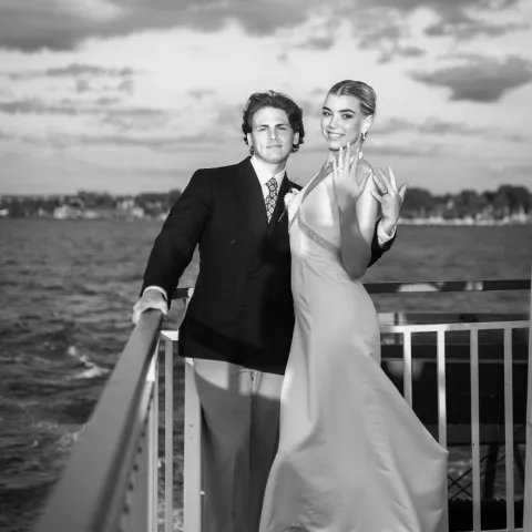Couple posing on a boat deck, smiling, with water and cloudy sky in the background.