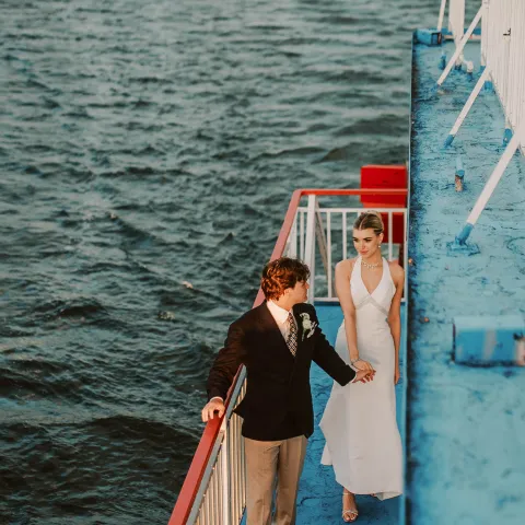Couple walking on a ship deck near the ocean, holding hands.