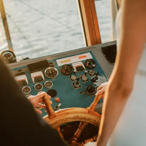 Two people steering a boat with a wooden wheel at sunset.