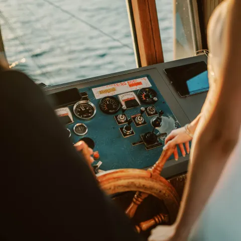 Two people steering a boat with visible control panel and water view.