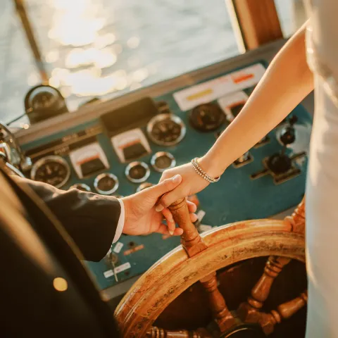 Couple holding hands on a ship's wheel at sunset.