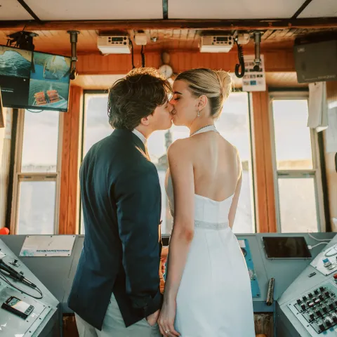 Couple in wedding attire standing close in a boat's control room.