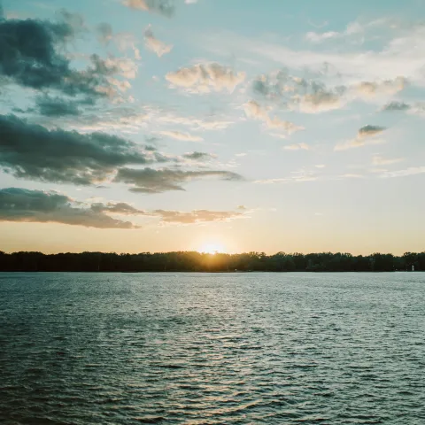 Sunset over a calm lake with scattered clouds and distant treeline.