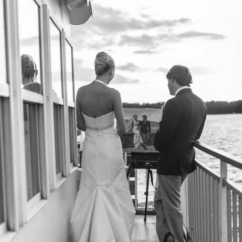 A couple on a boat deck, facing away, gazing at water under a cloudy sky.