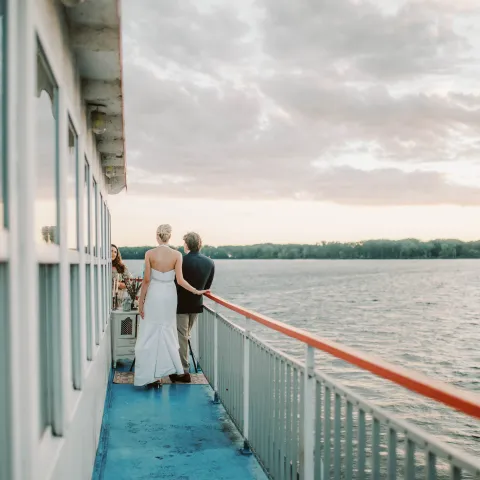 Couple in formal wear stands on boat deck overlooking calm water at sunset.