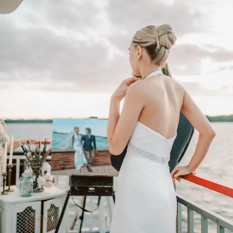 Bride in white dress looks at painting on a boat deck.