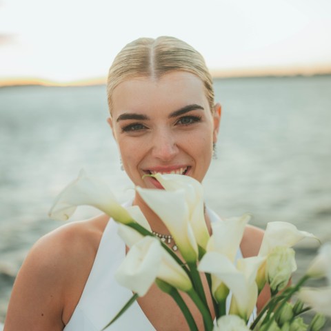 Woman smiling, holding white flowers by a lake at sunset.