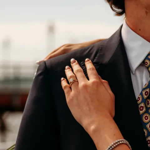 Person's hand with ring resting on a suited shoulder outdoors.