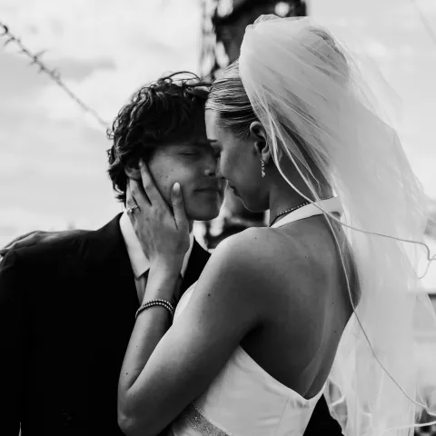 Bride and groom embrace, touching foreheads, against a sky background.