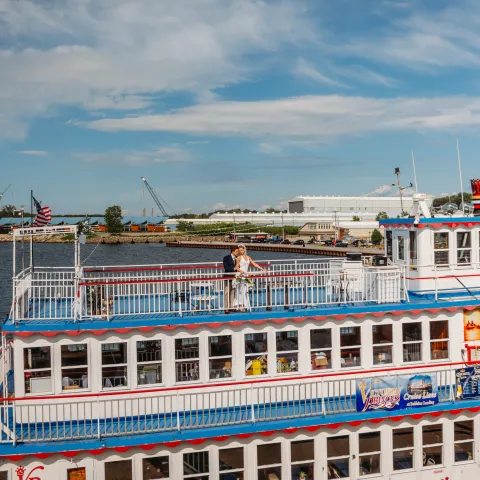 White and blue riverboat docked with people on top deck, under a partly cloudy sky.