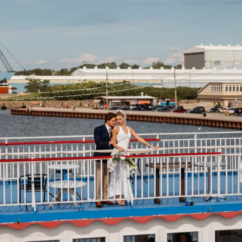 Couple in wedding attire on a blue and white boat by an industrial waterfront.