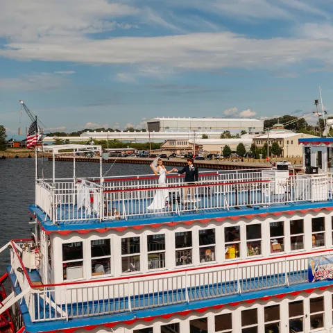 Riverboat with white railings and a couple on the deck under a blue sky.