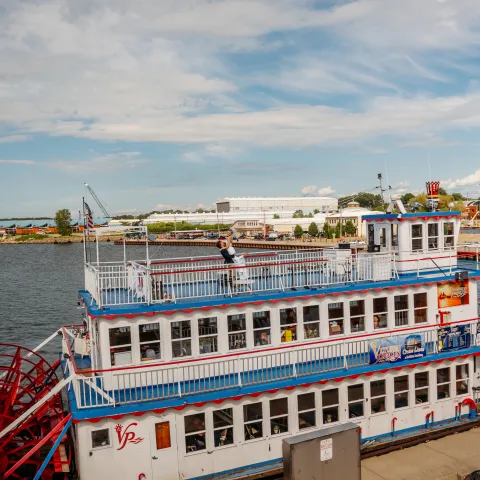 Riverboat with red paddle wheel docked under a partly cloudy blue sky.