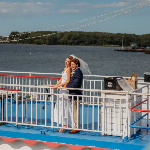 Couple in wedding attire on a boat deck overlooking water.