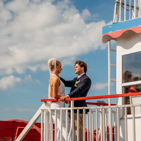 Couple in formal attire on a boat with blue sky and clouds.