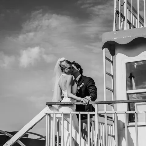 Bride and groom hugging on a boat deck under a cloudy sky.
