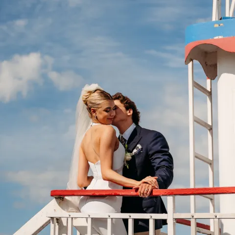 Bride and groom embrace on a boat against blue sky.