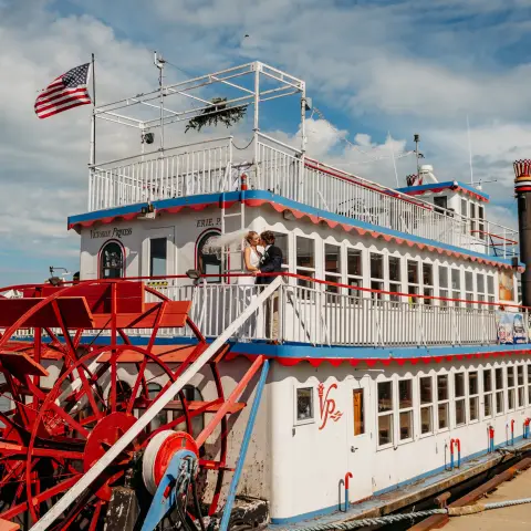 Steamboat with paddle wheel docked by a riverside, American flag on top, under a blue sky with clouds.