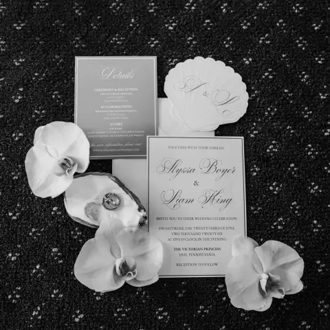 Black and white wedding invitations surrounded by white flowers on a textured surface.