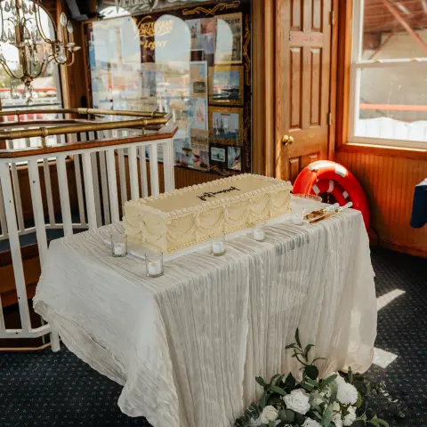 Cake on a table with white cloth and candles, surrounded by flowers in a carpeted room.