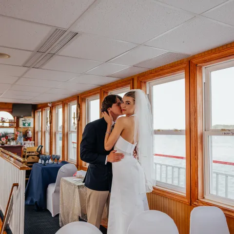 Bride and groom kissing on a boat, surrounded by decorated tables and windows.